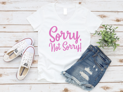 Flat lay of a white t-shirt with pink text that reads “Sorry. Not Sorry!”, styled with denim shorts, white Converse sneakers, and a small green plant on a white wooden background. www.missmafia.com