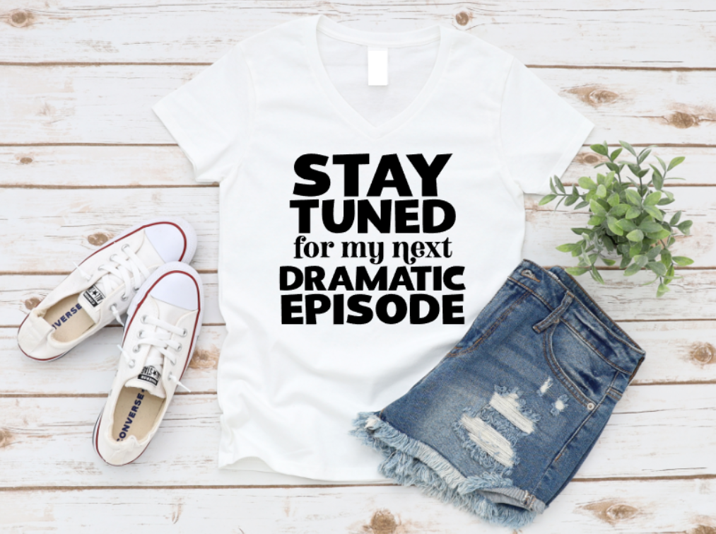Flat lay of a white t-shirt with the bold phrase “Stay Tuned for My Next Dramatic Episode” in white text, styled with distressed denim shorts, white Converse sneakers, and a potted green plant on a white wooden background. www.missmafia.com