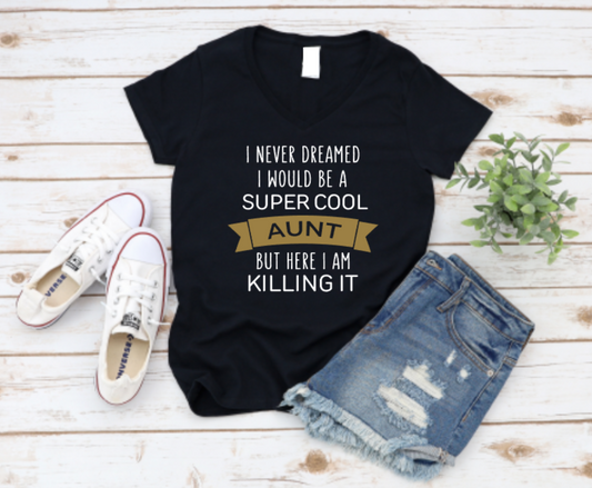 Black t-shirt with “I never dreamed I would be a super cool aunt but here I am killing it” text in white and gold, styled with white Converse sneakers and distressed denim shorts on a light wooden background.