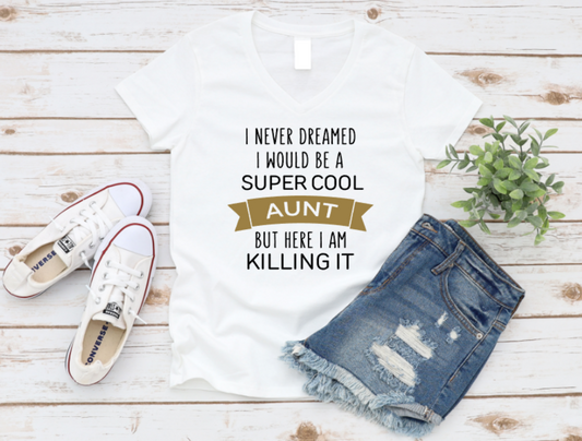 White t-shirt with “I never dreamed I would be a super cool aunt but here I am killing it” text in white and gold, styled with white Converse sneakers and distressed denim shorts on a light wooden background.
