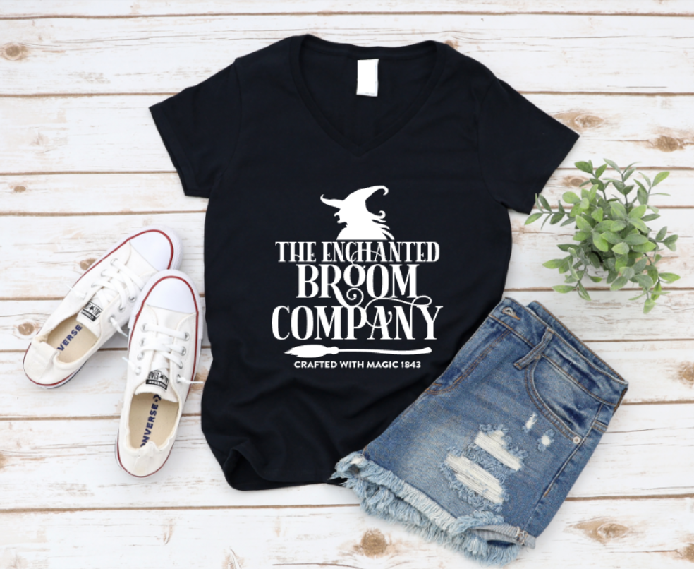 Black t-shirt with 'The Enchanted Broom Company' design on a wooden surface with shoes and shorts. www.MissMafia.com