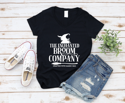 Black t-shirt with 'The Enchanted Broom Company' design on a wooden surface with shoes and shorts. www.MissMafia.com