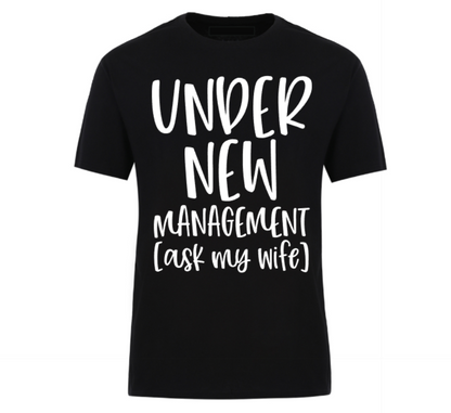 Black t-shirt with bold black text reading “Under New Management – See Wife for Details,” styled with casual denim and sneakers—funny gift for husbands or newlyweds.