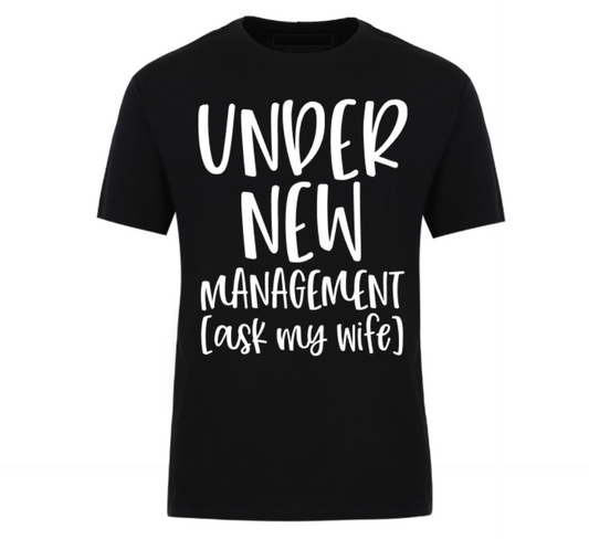 Black t-shirt with bold black text reading “Under New Management – See Wife for Details,” styled with casual denim and sneakers—funny gift for husbands or newlyweds.