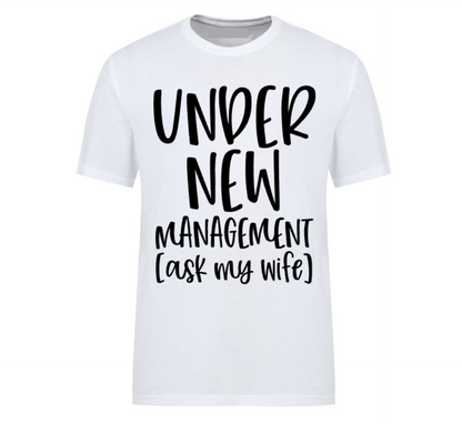 White t-shirt with bold black text reading “Under New Management – See Wife for Details,” styled with casual denim and sneakers—funny gift for husbands or newlyweds.