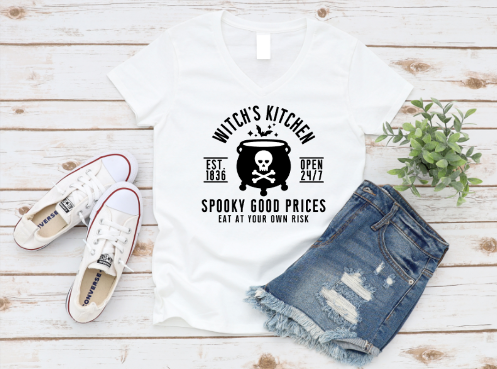 Flat lay of a white t-shirt with a white Halloween-themed design that reads “Witch’s Kitchen – Spooky Good Prices – Eat At Your Own Risk,” featuring a bubbling cauldron with a skull and crossbones. Styled with distressed denim shorts, white Converse sneakers, and a small green plant on a white wood background. www.missmafia.com